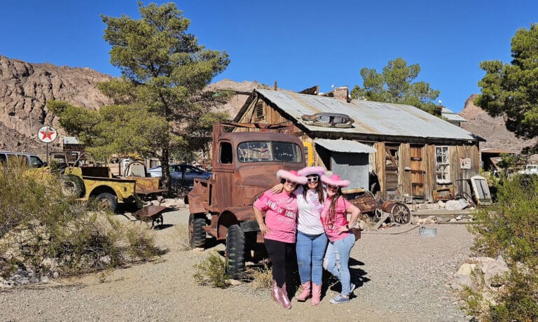 Three in pink cowboy attire pose by a rusty truck in a Nevada ghost town near Las Vegas, with vintage cars and desert hills.
