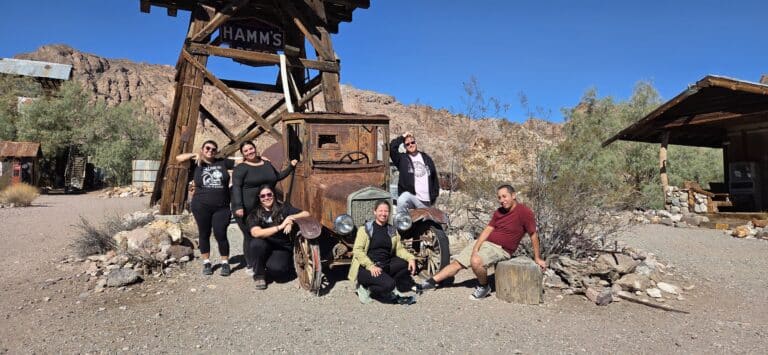 Group of six by a rusty vintage car in a Nevada desert ghost town near HAMMS sign, rocky hills, and cabins close to Las Vegas.