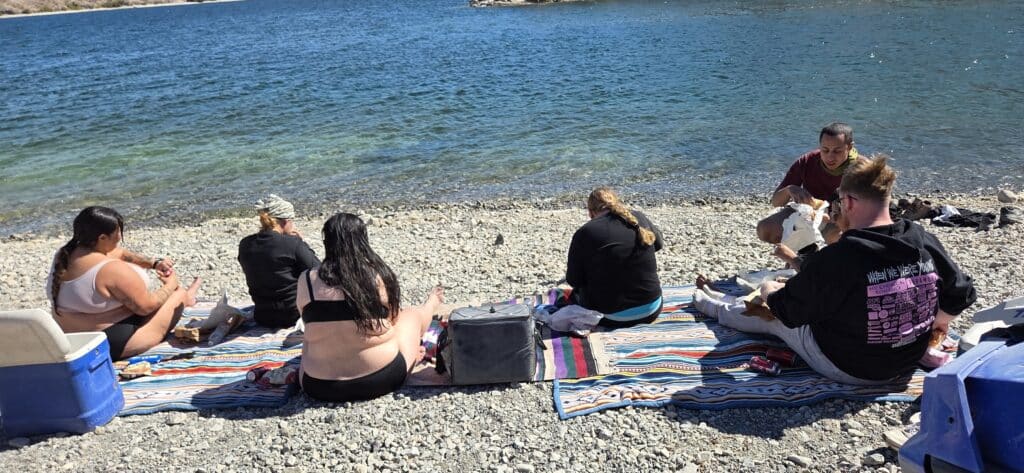 Friends picnic on colorful blankets by the Colorado River near Las Vegas, Nevada, enjoying drinks under bright sun.