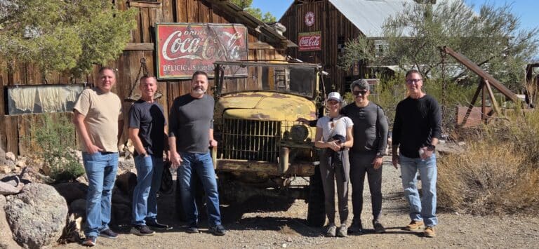 Group poses by a rustic wooden building and vintage yellow truck in a Nevada ghost town near the Colorado River.