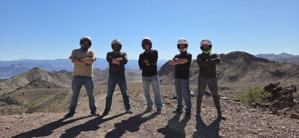 Group in helmets and sunglasses poses on Nevada rocky overlook, with blue sky, during Las Vegas ATV tour near Colorado River.