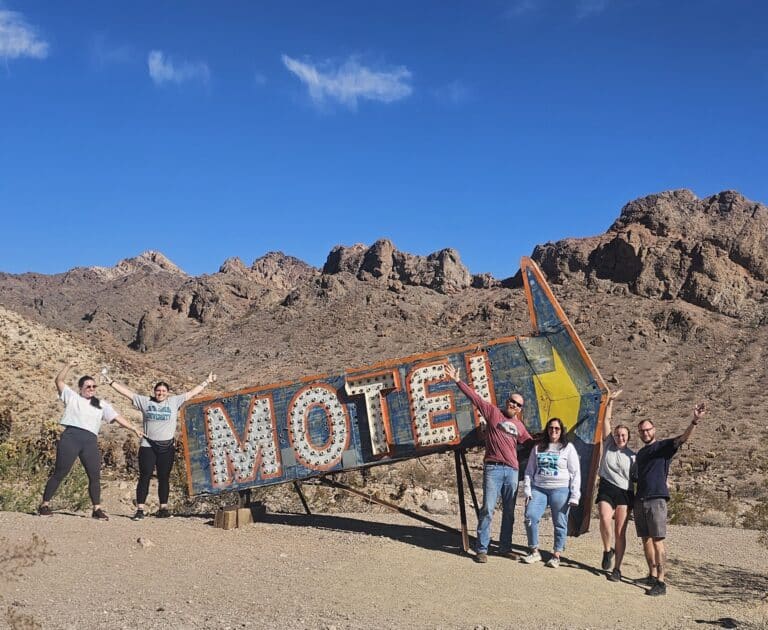 Group smiles by vintage arrow-shaped MOTEL sign near Nevada desert, with rocky mountains and clear sky—perfect for Las Vegas road trips.