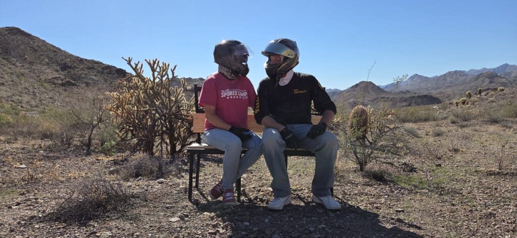Two people in helmets hold hands on a desert bench near Las Vegas, Nevada, ready for ATV tours with mountains and cacti behind.