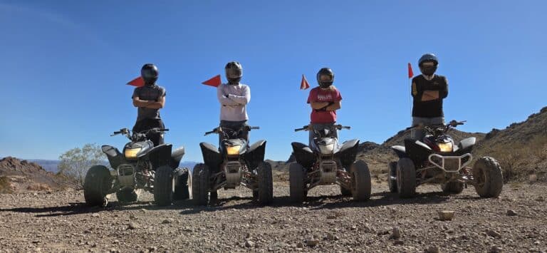 Group in helmets pose by ATVs with red flags on a rocky Nevada desert ATV tour near Las Vegas under clear blue sky.