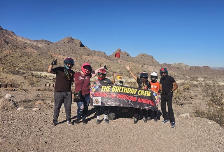 Six helmeted riders in Nevada’s desert hold a “Birthday Crew” banner on an epic ATV tour near Las Vegas under blue skies.