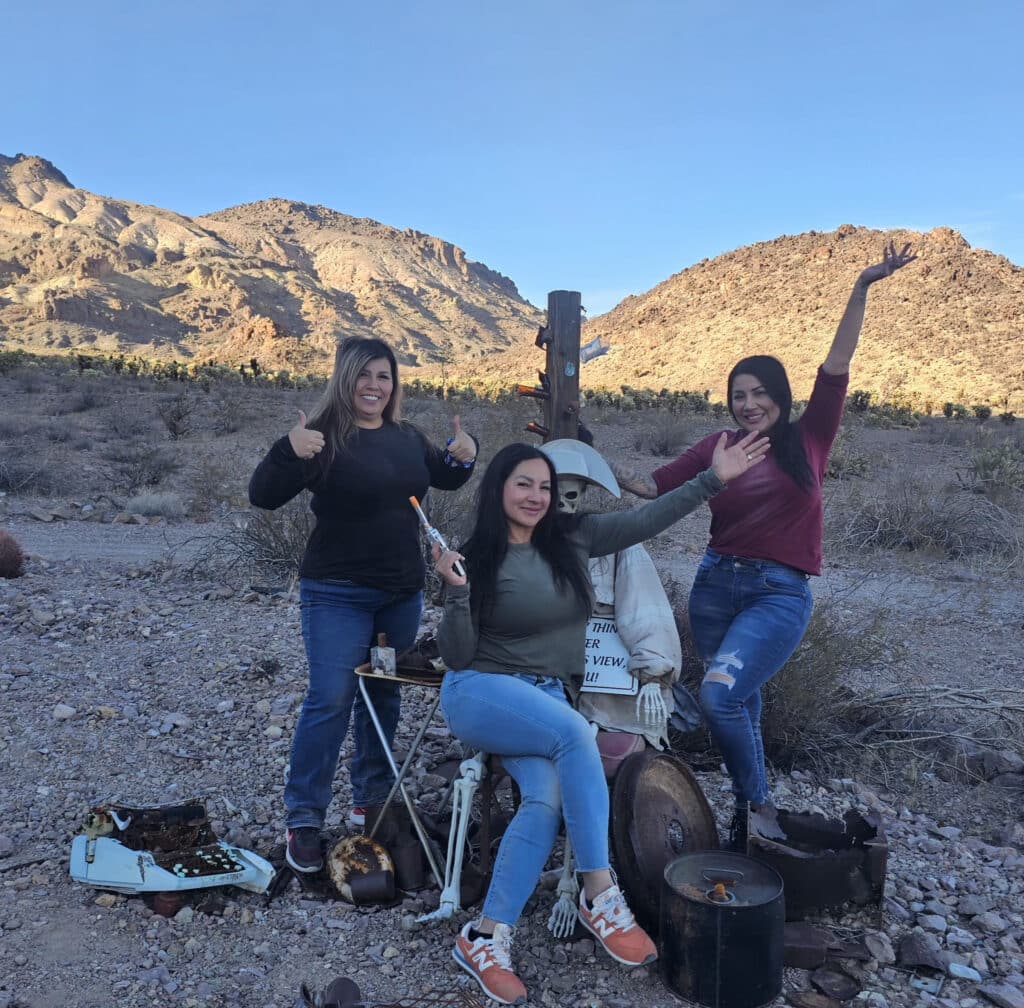 Three women pose by a prospector scarecrow in a Nevada desert near rocky hills—perfect for Ghost Town sightseeing in Las Vegas.