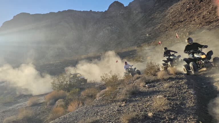 Adventurers ride ATVs on a dusty Nevada desert trail, near Las Vegas, with mountains and blue sky in the distance.