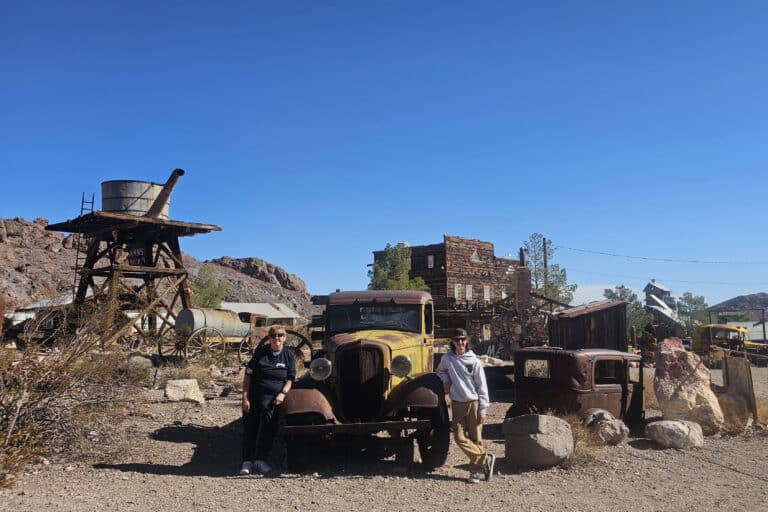 Tourists by a rusty antique car in a Nevada desert ghost town near the Colorado River, ideal for Las Vegas ghost town sightseeing.