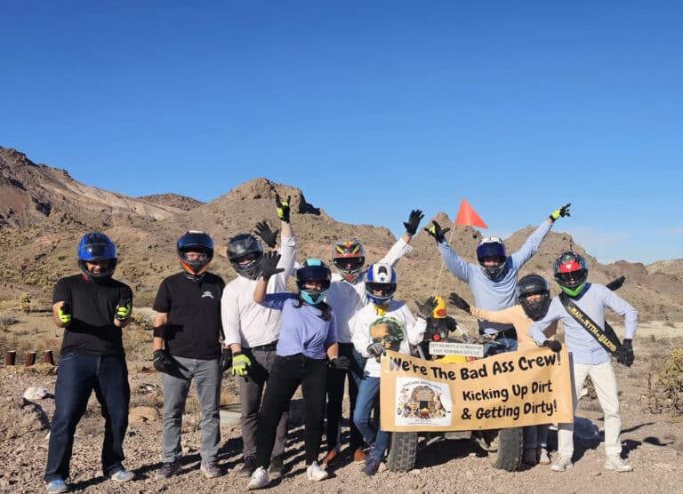 Group in helmets poses in Nevada desert near Las Vegas after ATV tour, holding banner; mountains and Colorado River visible.