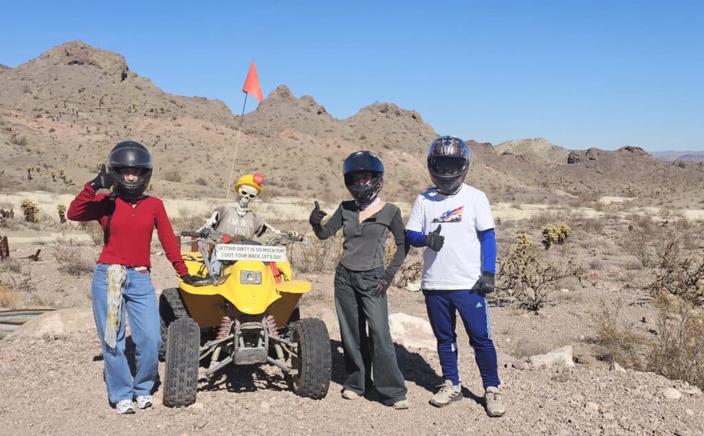 ATV tour near Las Vegas: three helmeted riders pose by a skeleton on a quad in the Nevada desert, mountains and cacti behind.