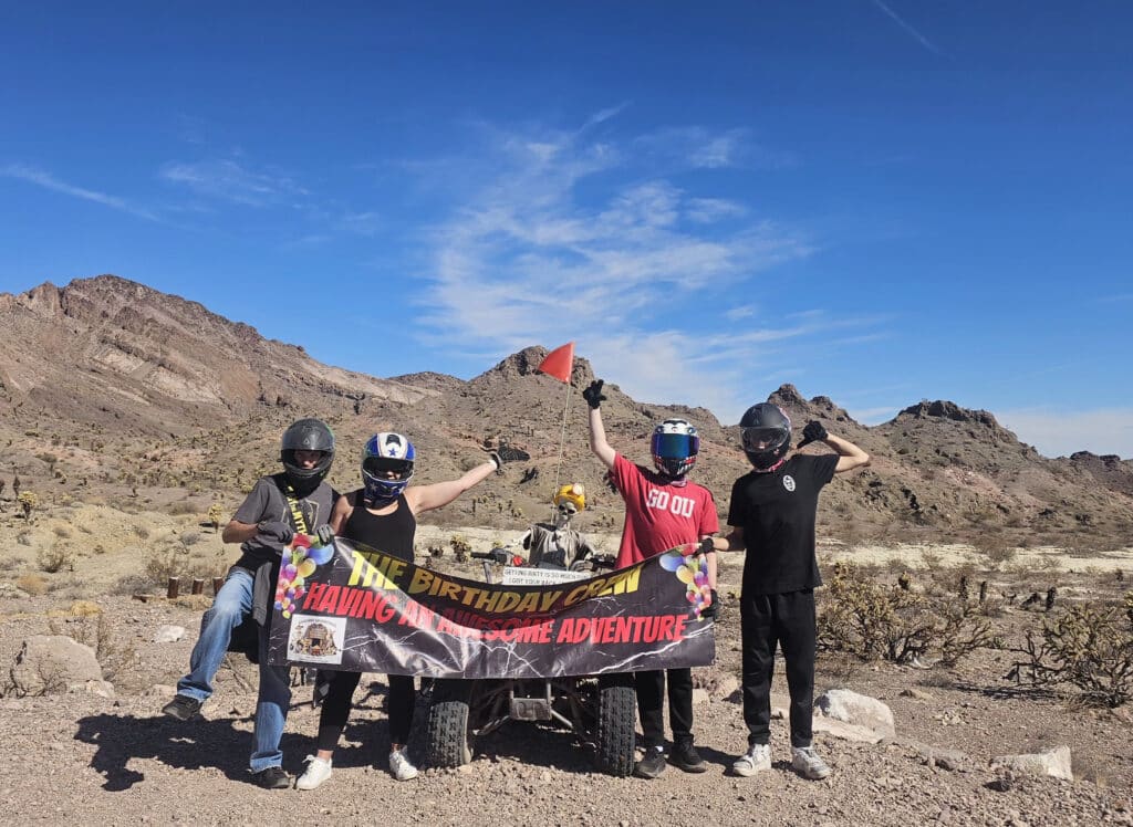 Group in helmets holds a birthday banner by an ATV near Las Vegas, Nevada, with desert views and rocky mountains under blue sky.