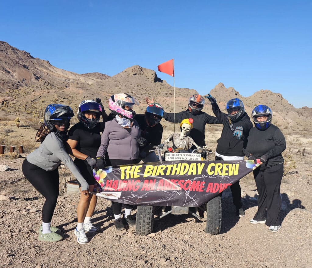 Group in helmets pose with quad bike and "The Birthday Crew" banner in Nevada desert, ready for Las Vegas ATV tour adventure.