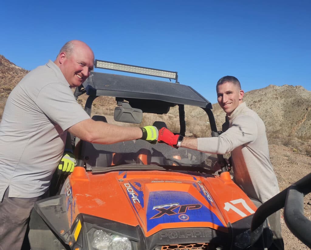 Two men enjoy a RZR off-road ride near Las Vegas, Nevada, fist bumping by their orange ATV in the desert under blue skies.