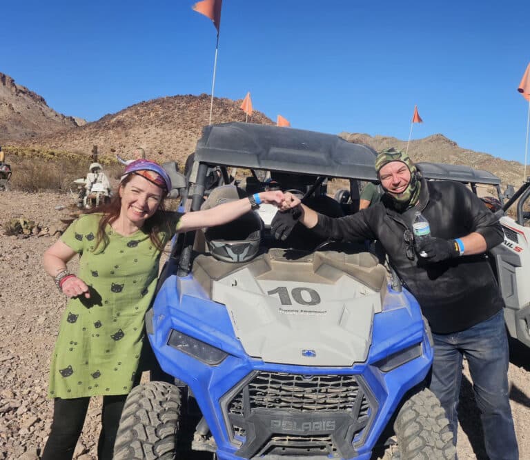 Smiling couple holds hands by RZR #10 on a Nevada desert ATV tour near Las Vegas; orange flags wave, rocky landscape behind.