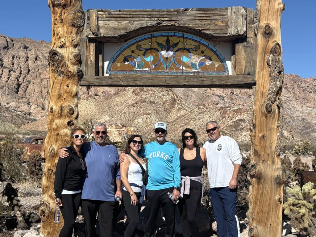 Group poses by a rustic wooden arch with stained glass in Nevada desert near Las Vegas, cacti and rocky hills under blue sky.