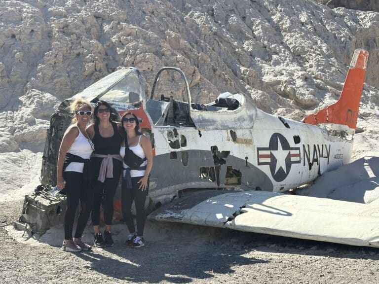 Three women in activewear smile by an old Navy plane in a Nevada desert near Las Vegas, ideal for ATV tours and ghost town sightseeing.