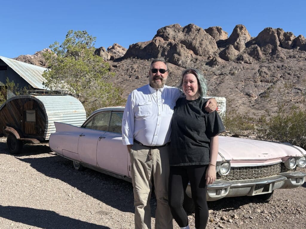 Couple smiles by vintage pink Cadillac near Nevada ghost town, with rocky desert, clear sky—perfect for Las Vegas sightseeing.