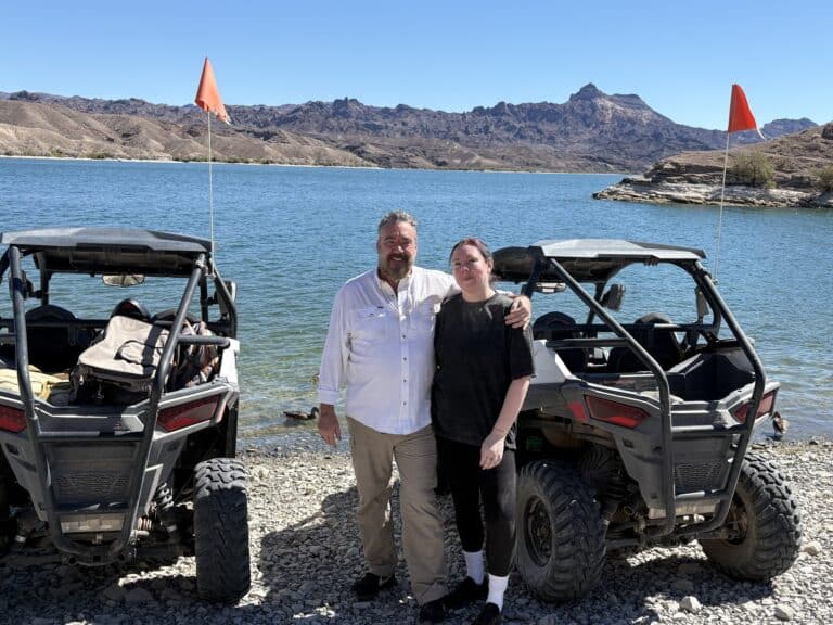 Smiling pair by two ATVs on rocky Colorado River lakeshore near Las Vegas, mountains and blue water under Nevada sky.