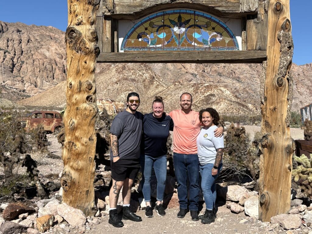 Smiling group under rustic arch near cacti in Nevada desert, with old car—perfect for Las Vegas Ghost Town sightseeing tours.
