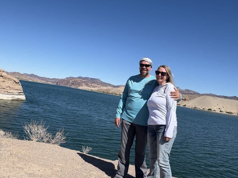 Smiling couple in sunglasses by Colorado River near Las Vegas, Nevada, enjoying a sunny day outdoors with rocky hills in view.