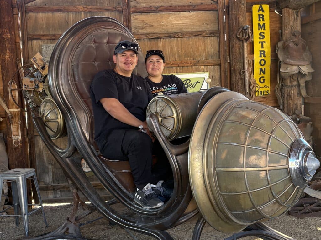 Two people on a steampunk chair in Nelson Ghost Town near Las Vegas, Nevada, with saddles and an Armstrong Tires sign behind.