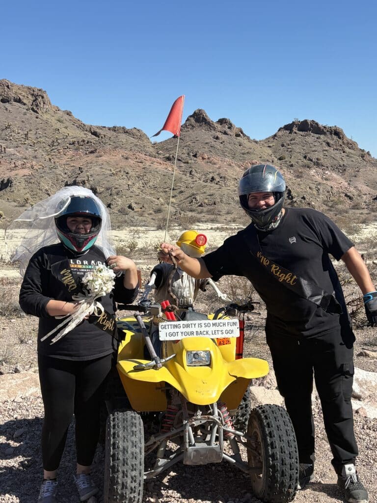 Two people in helmets hold hands and a bird by a yellow ATV on a Nevada desert ATV tour near Las Vegas with mountains behind.