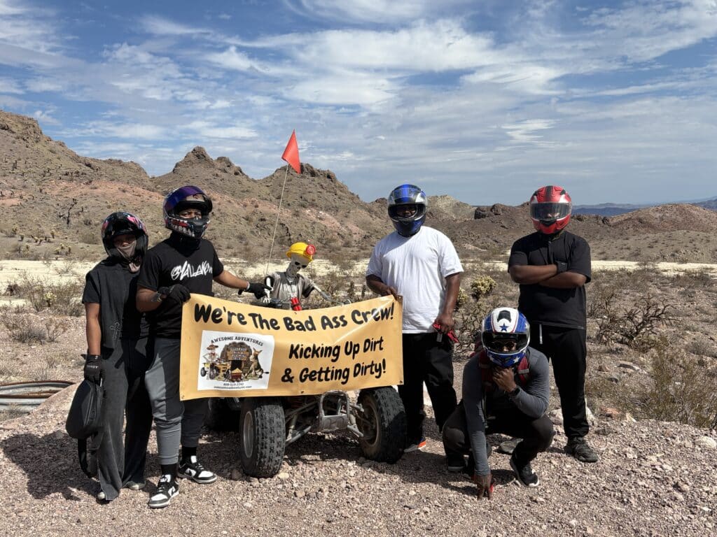 Group in helmets with ATV poses in Nevada desert, banner reads "We’re The Bad Ass Crew!" Perfect for Las Vegas ATV tours.