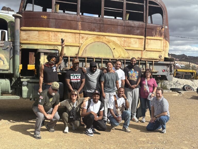 Thirteen people posing by a rusted bus in Nevada desert near Las Vegas, enjoying a Ghost Town sightseeing adventure.