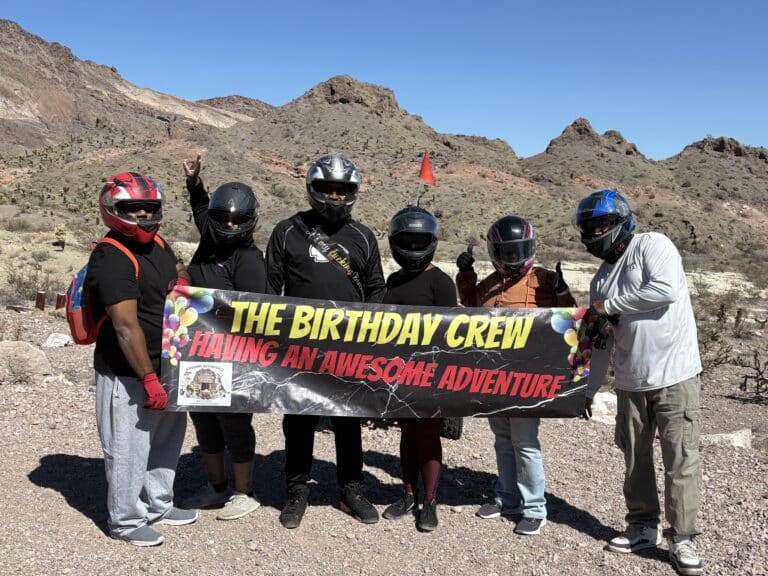 Group in helmets with banner “THE BIRTHDAY CREW HAVING AN AWESOME ADVENTURE” on rocky Nevada terrain, near Colorado River.