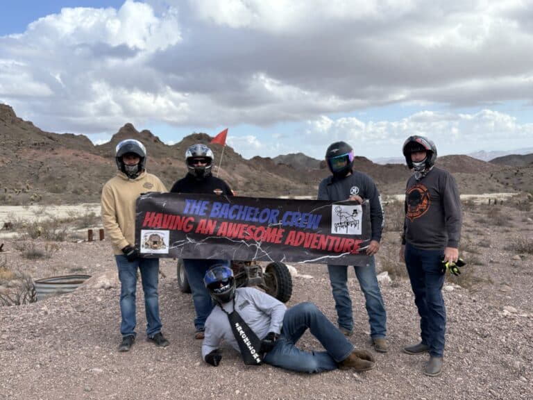 Group in helmets holds 'The Bachelor Crew Having an Awesome Adventure' banner on a Nevada ATV tour near Las Vegas rocky desert.