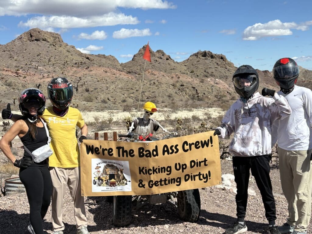 Group in helmets and gear holds "Bad Ass Crew" sign on a Nevada desert ATV tour near Las Vegas under clear blue sky.
