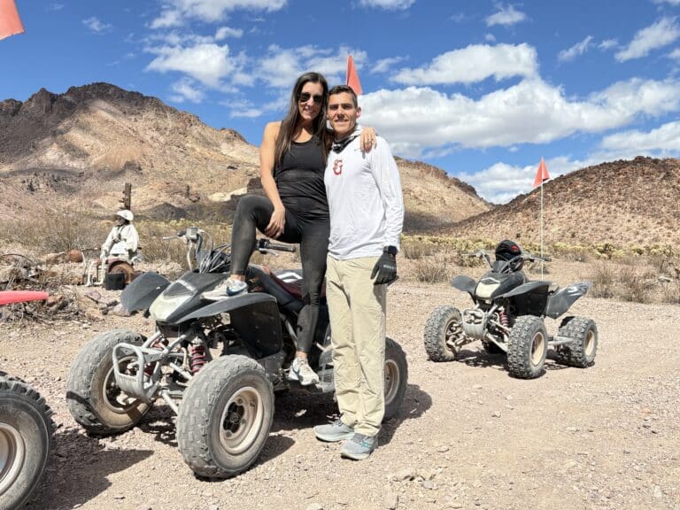 Couple smiles on an ATV during a Nevada desert RZR tour near Las Vegas, with mountains, blue sky, and another ATV in the background.