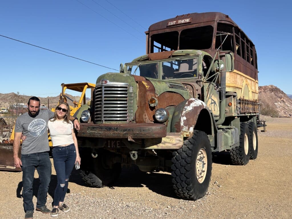 Smiling couple by a rusty off-road bus near Nevada desert, perfect for Las Vegas ATV tours or Ghost Town sightseeing.