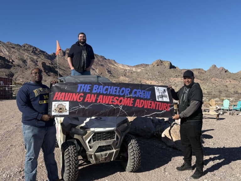 Three men in Nevada pose with a "Bachelor Crew" banner before rocky hills—Las Vegas RZR off-road adventure under blue sky.