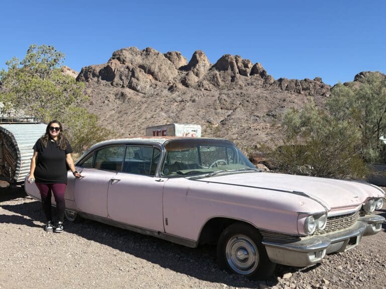 A woman in sunglasses stands by a vintage pink car in the Nevada desert near Las Vegas, with mountain views and blue skies.