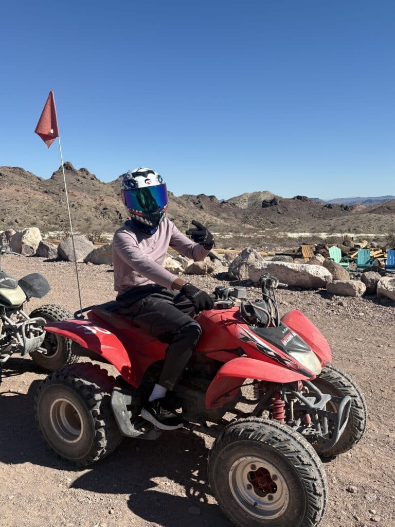 Rider on red ATV giving thumbs-up in Nevada desert near Las Vegas with mountains, red flag, and blue sky—perfect for ATV tours.