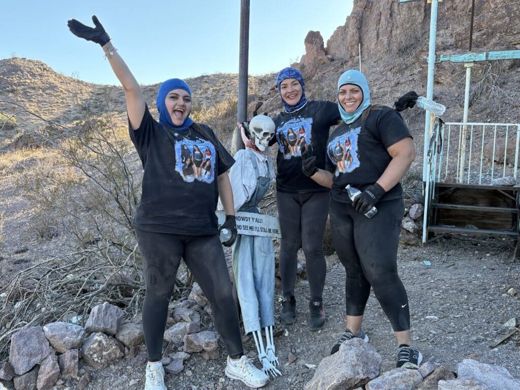 Three women in black shirts and blue headscarves pose with a skeleton prop in the Nevada desert near Las Vegas Ghost Town.