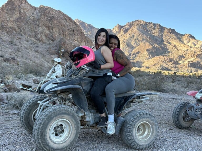 Two women enjoy a Las Vegas ATV tour in a rocky Nevada desert, mountains behind, pink helmet in hand, another rider nearby.