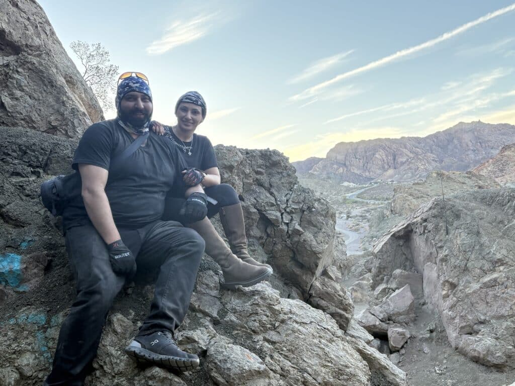 Two hikers in black gear smile on Nevada’s rocky terrain at sunset, mountains and Colorado River near Las Vegas in view.