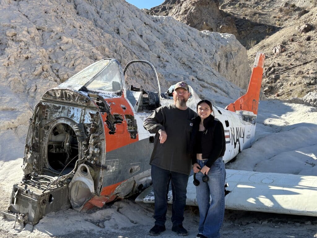 Smiling duo by a vintage Navy plane wreck on a sunny Nevada desert, perfect for Las Vegas ATV tours or Ghost Town adventures.