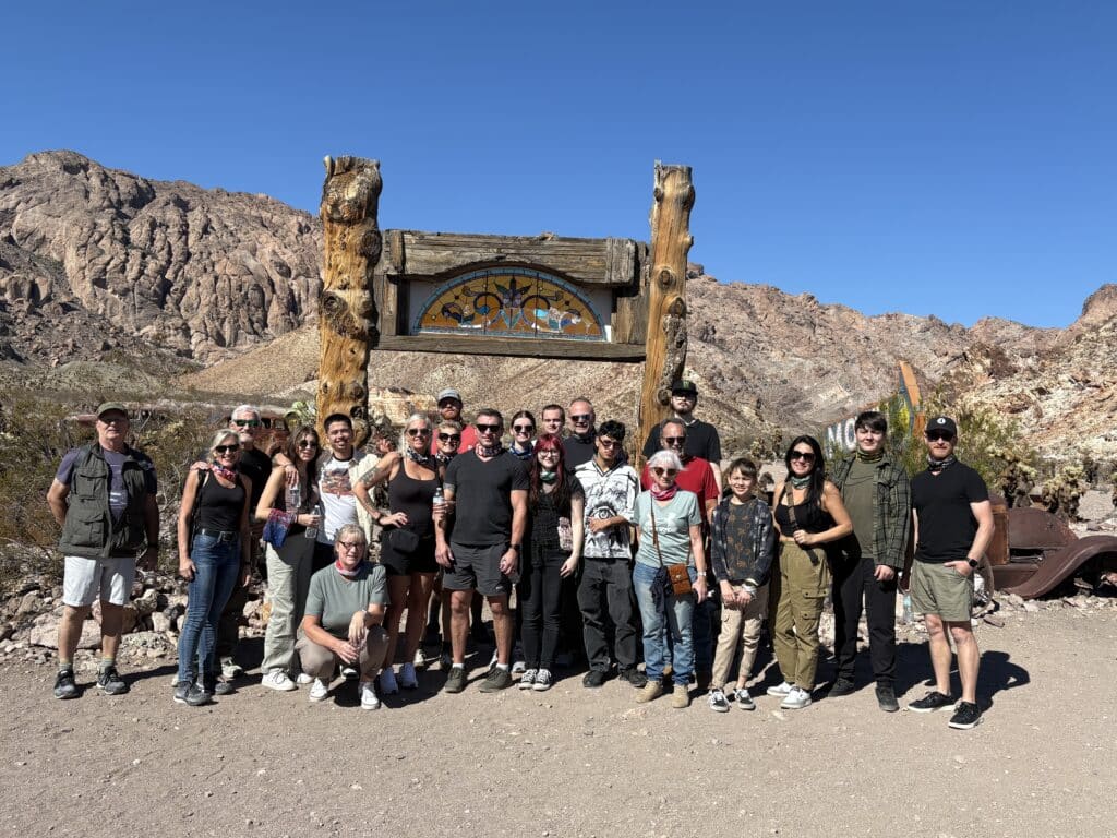 Group of 20 smiling in front of rustic sign near Nevada mountains, perfect for Las Vegas ATV tours or Ghost Town sightseeing.