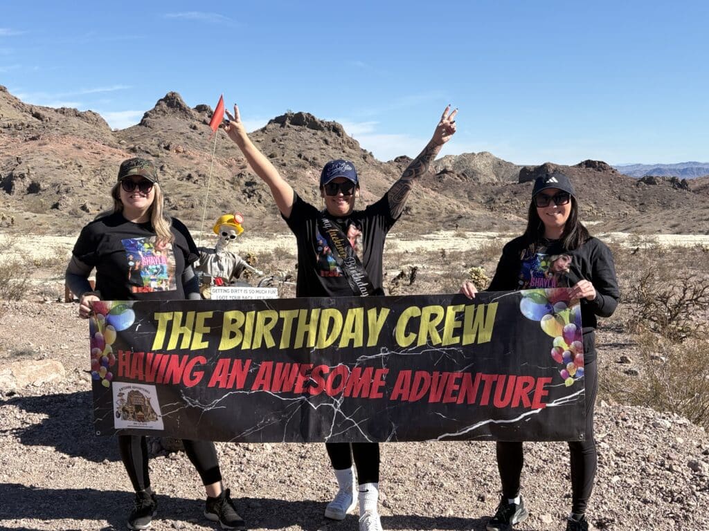 Three women in the Nevada desert near Las Vegas smile, holding a “Birthday Crew Adventure” banner by rocky hills and blue sky.