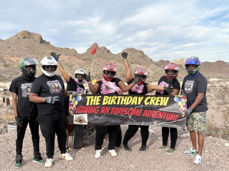 Six friends in helmets on Nevada rocky terrain hold a "Birthday Crew" banner, ready for a Las Vegas ATV adventure tour.