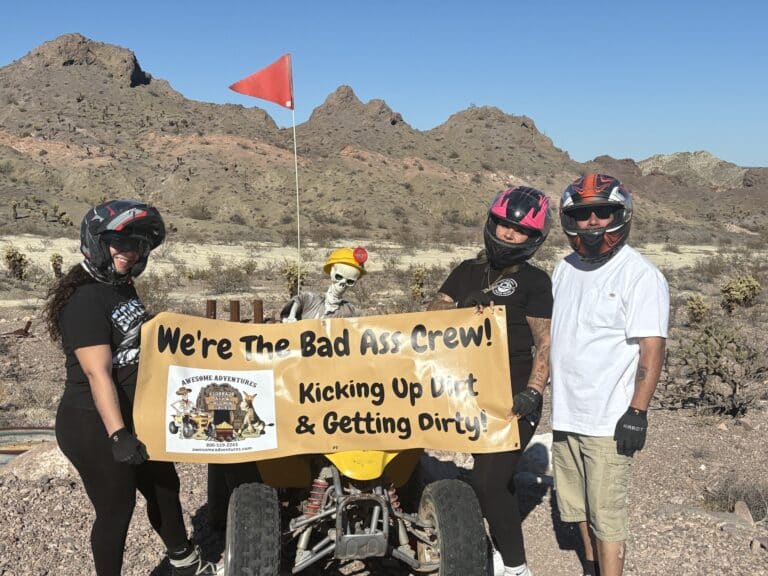ATV riders in helmets pose near a skeleton on an ATV in the Nevada desert, holding a "Bad Ass Crew" banner during an off-road tour.
