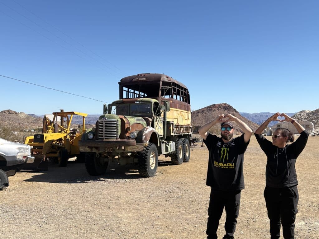 Two people make heart shapes on gravel in Nevada near an old truck, ATVs, and rocky hills, perfect for Las Vegas ghost town tours.
