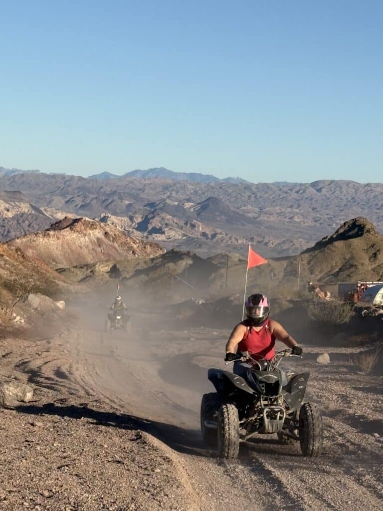 ATV tour near Las Vegas: two riders on a dusty Nevada trail with rocky mountains, blue sky, red flag, and safety gear.