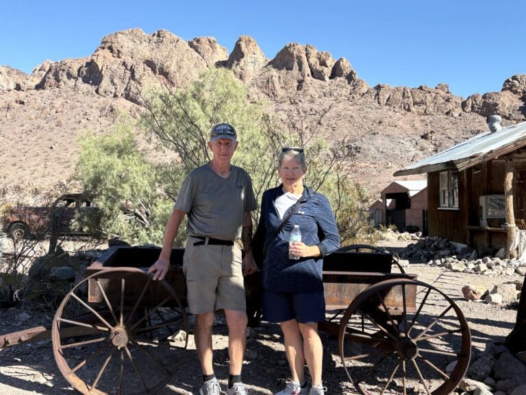 Two seniors by a wooden wagon in Nevada desert near Las Vegas, with Ghost Town sights and rocky hills under clear blue sky.