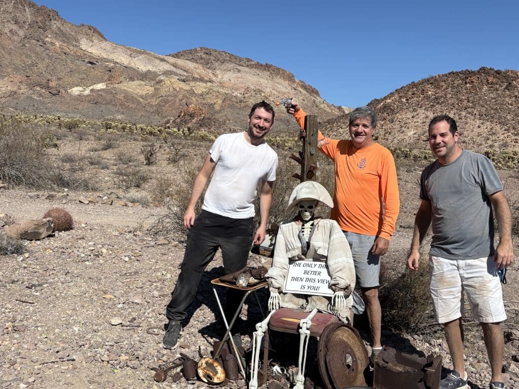 Three men in Nevada’s desert smile by a dressed skeleton holding a sign during a Las Vegas ATV tour near the Colorado River.