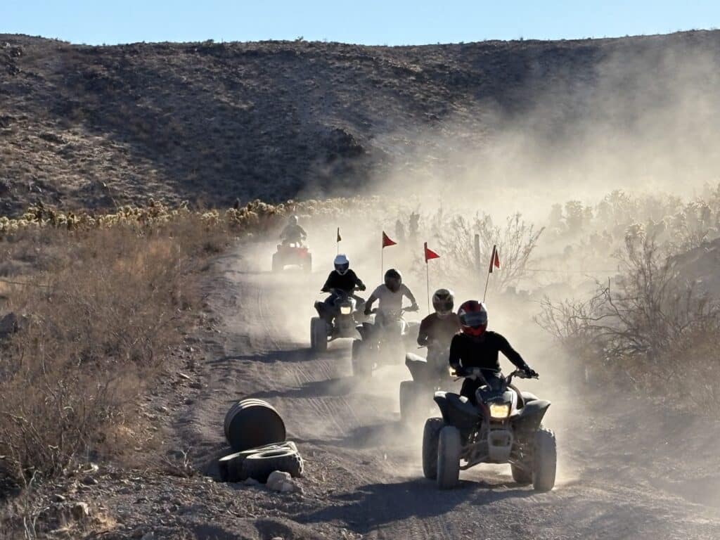 ATV riders in helmets speed along a dusty Nevada desert trail near Las Vegas, with red flags and scenic hills in the background.