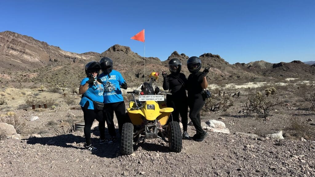 Riders in helmets pose by a yellow ATV on a Nevada desert tour near Las Vegas, with blue sky and mountains in the background.